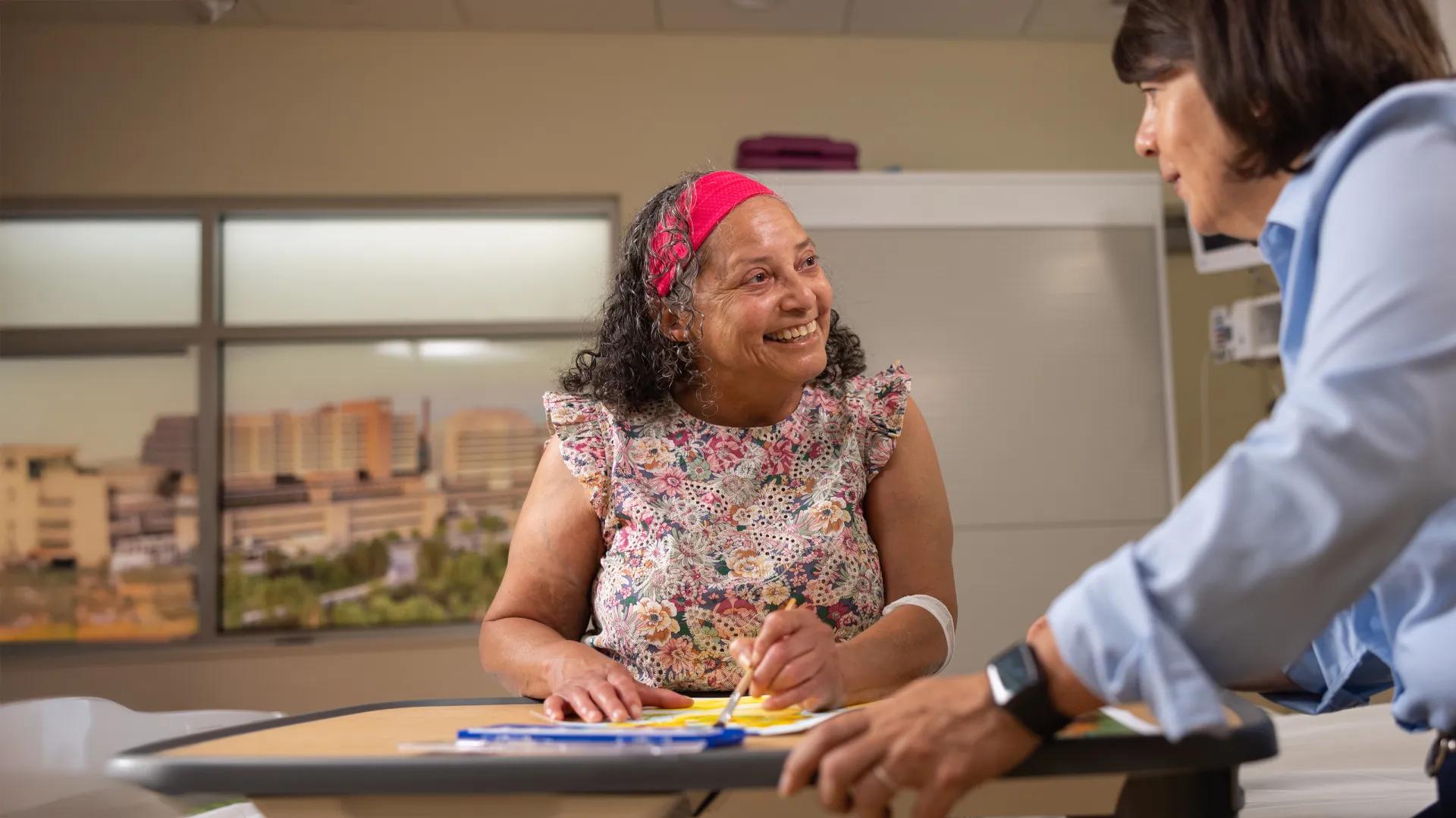 Female patient seated at table working on art project, smiling at woman looking on