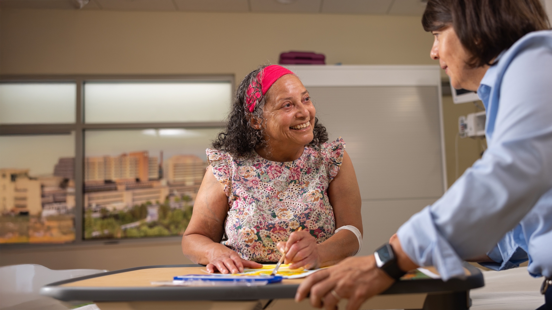 Female patient seated at table working on art project, smiling at woman looking on