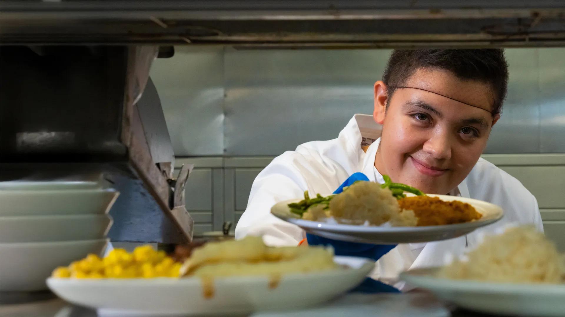 Smiling male food service worker wearing a hair net holding out a plate in a cafeteria setting