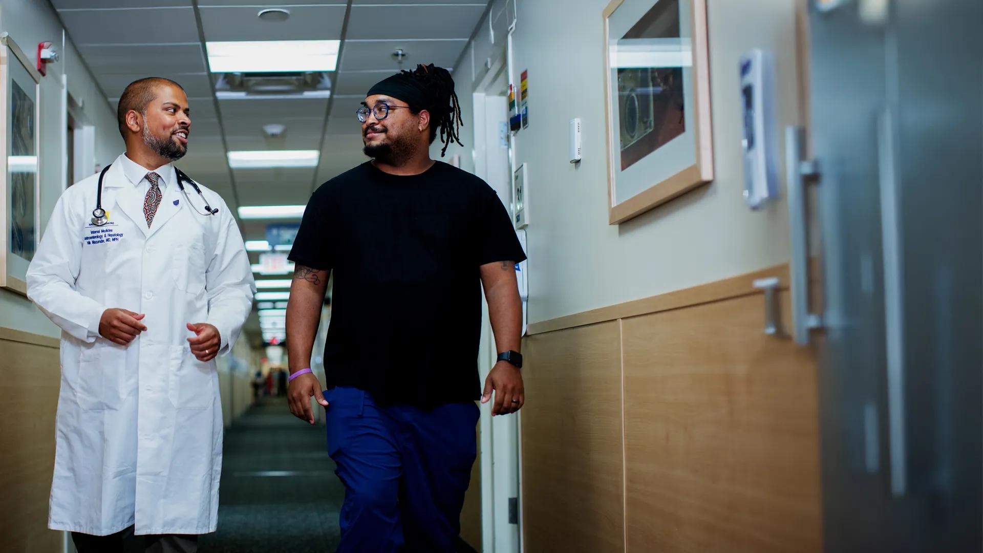 Black male doctor and black male with beard wearing black shirt and glasses walking and talking in hospital hallway
