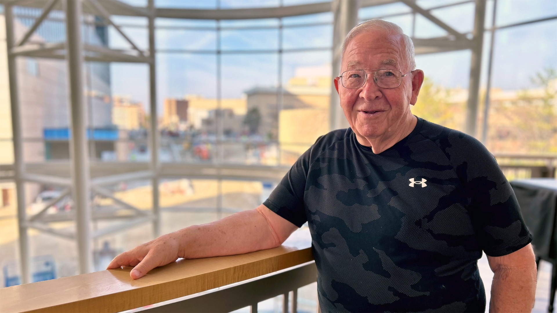 Older man wearing black camo t-shirt standing in Frankel Cardiovascular Center atrium with medical campus behind him