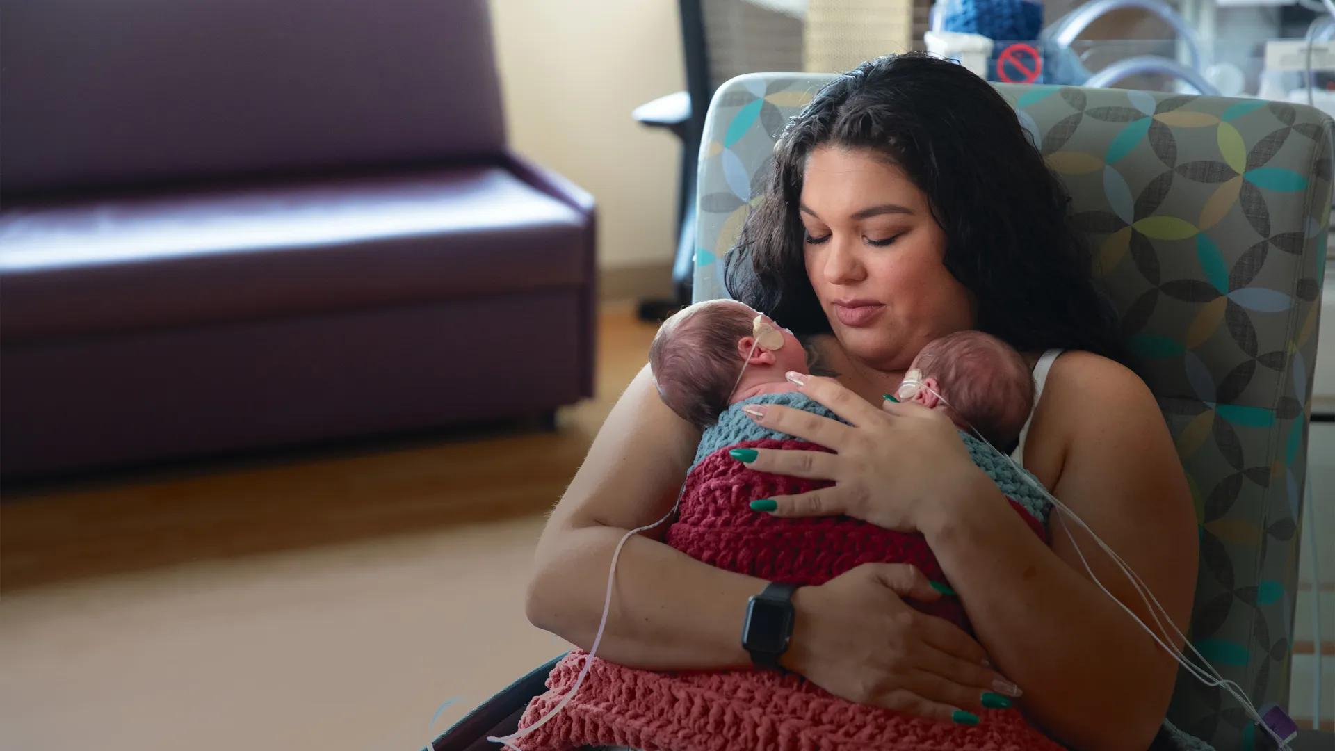 Mother seated in chair looking down at two tiny babies with medical tubes attached