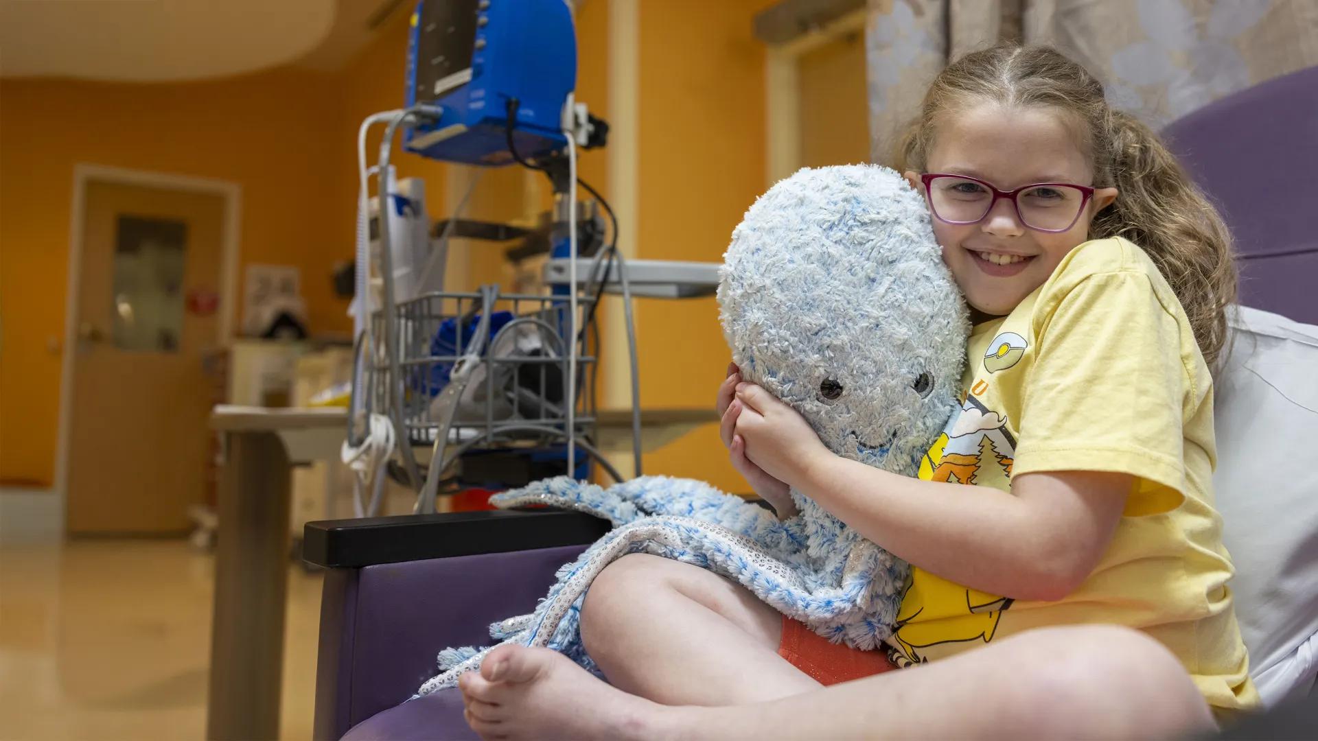 Girl with glasses in hospital bed hugging large stuffed octopus