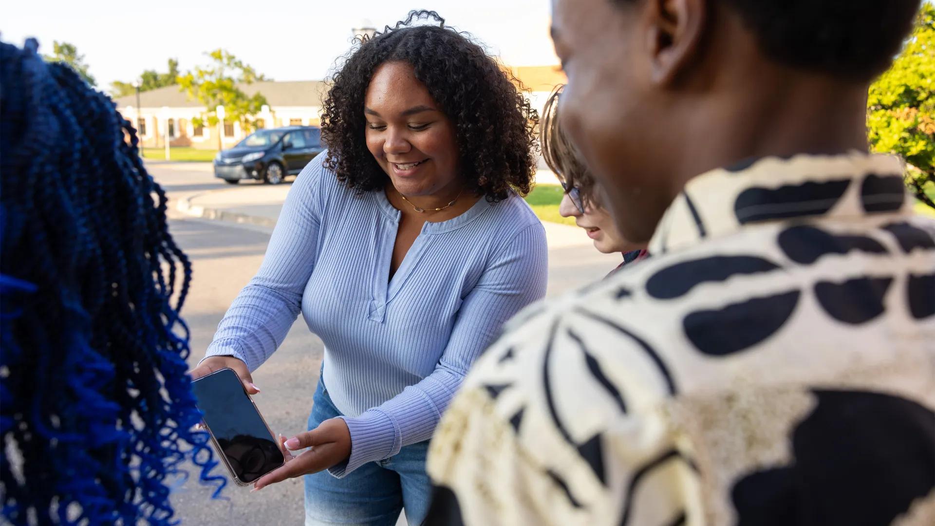 Young black woman standing in parking lot showing phone to group of diverse young adults