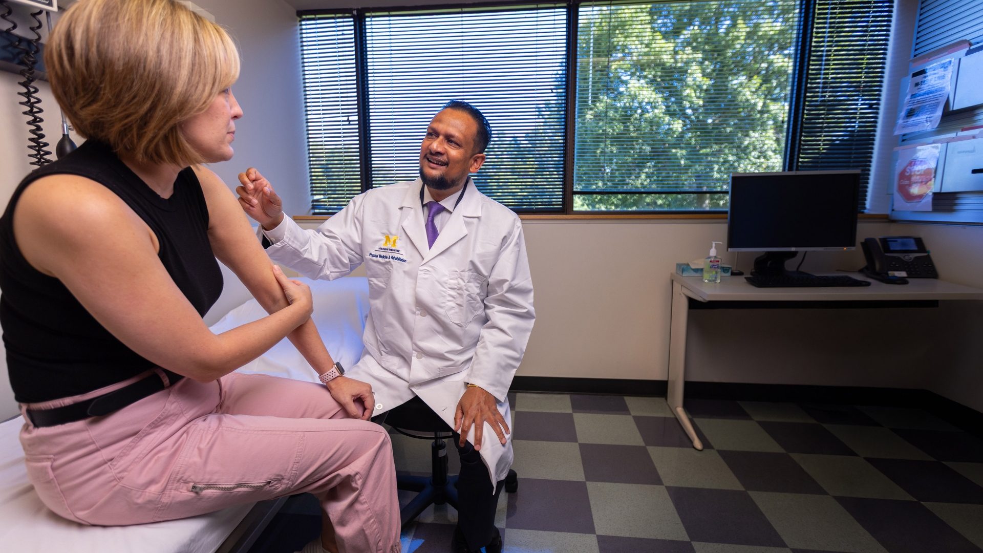 Doctor seated talking with female patient wearing pink pants and holding her arm 