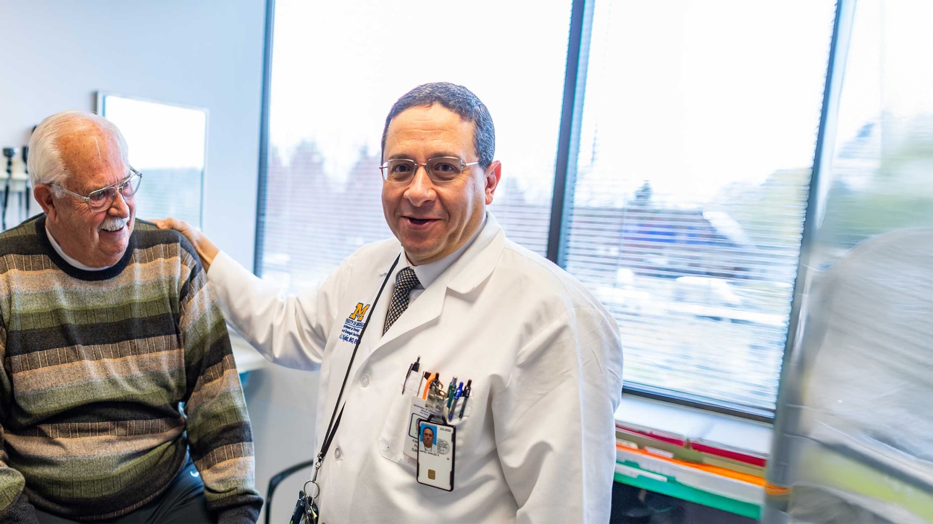 Smiling doctor with hand on shoulder of older male patient with a mustache wearing glasses and a striped shirt