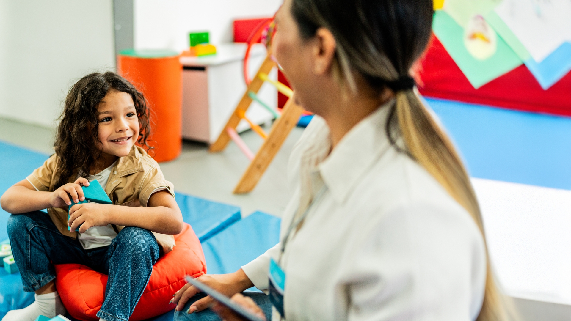 Smiling child seated on an orange cushion on a blue mat in a playroom, with a female health care provider looking on