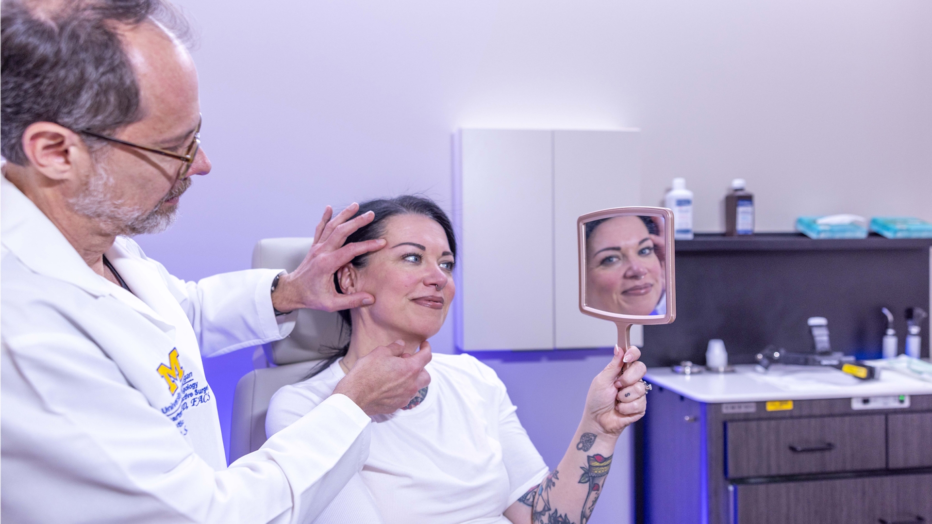Male doctor with glasses treating face of smiling female patient holding mirror