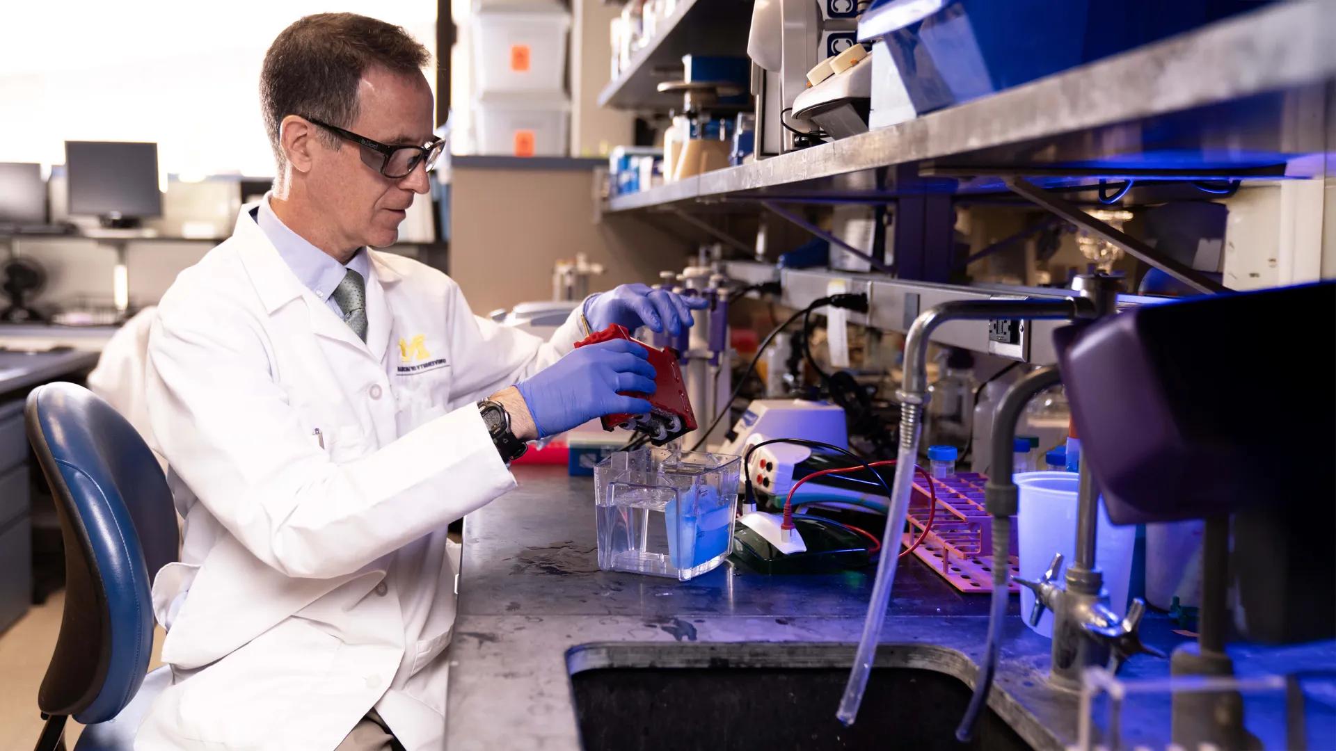 Man in white coat wearing lab goggles seated in lab in front of scientific equipment