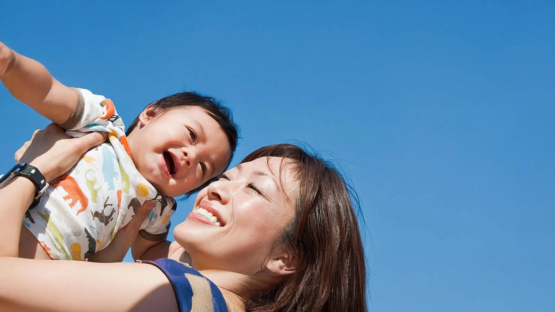 Smiling mother lifting a toddler in an animal print shirt against a bright blue sky  笑顔の母親が、明るい青空を背景にアニマル柄のシャツを着た幼児を持ち上げている様子