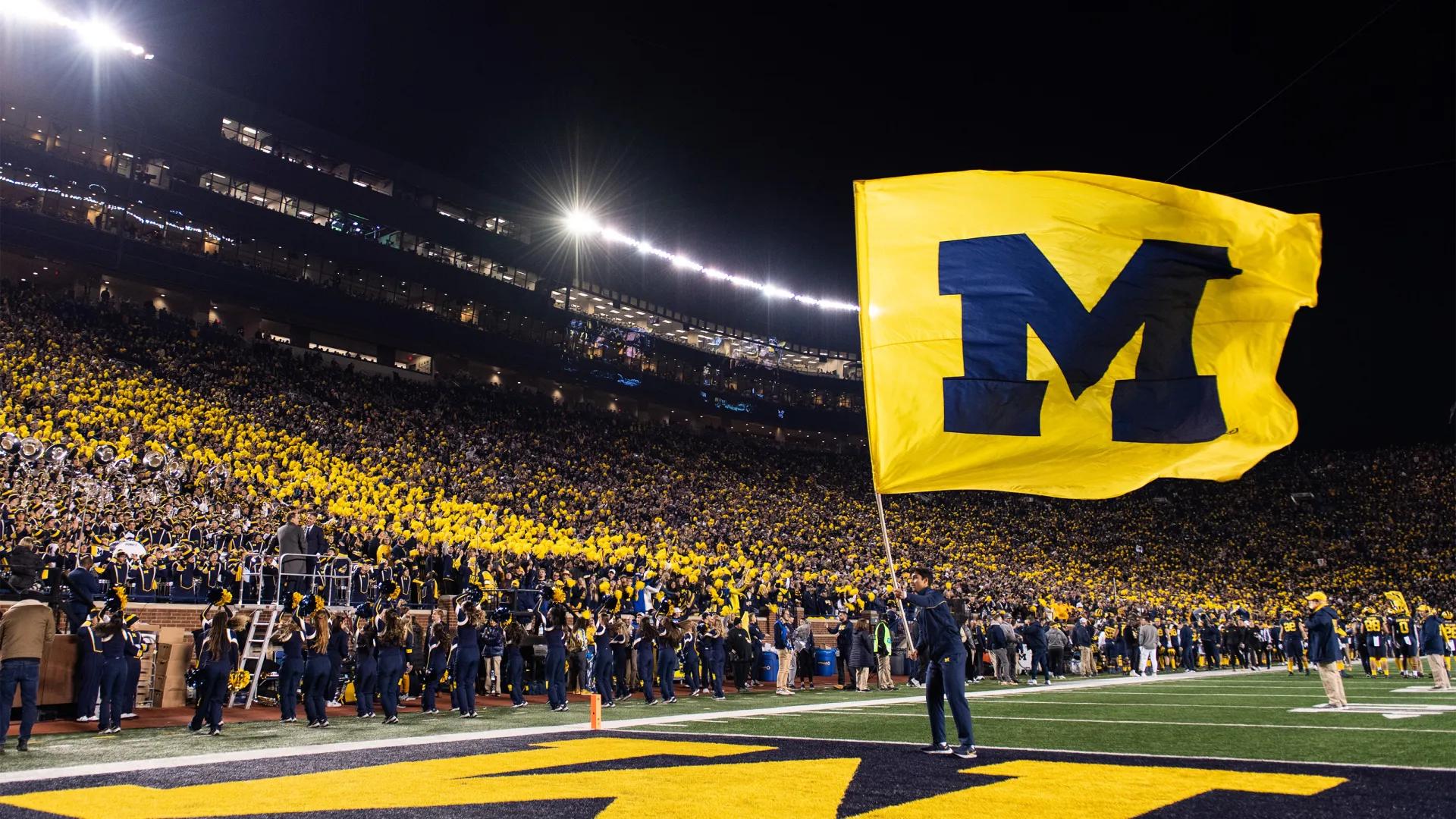 Man holding huge maize flag with blue block M at U-M football stadium with crowd in background