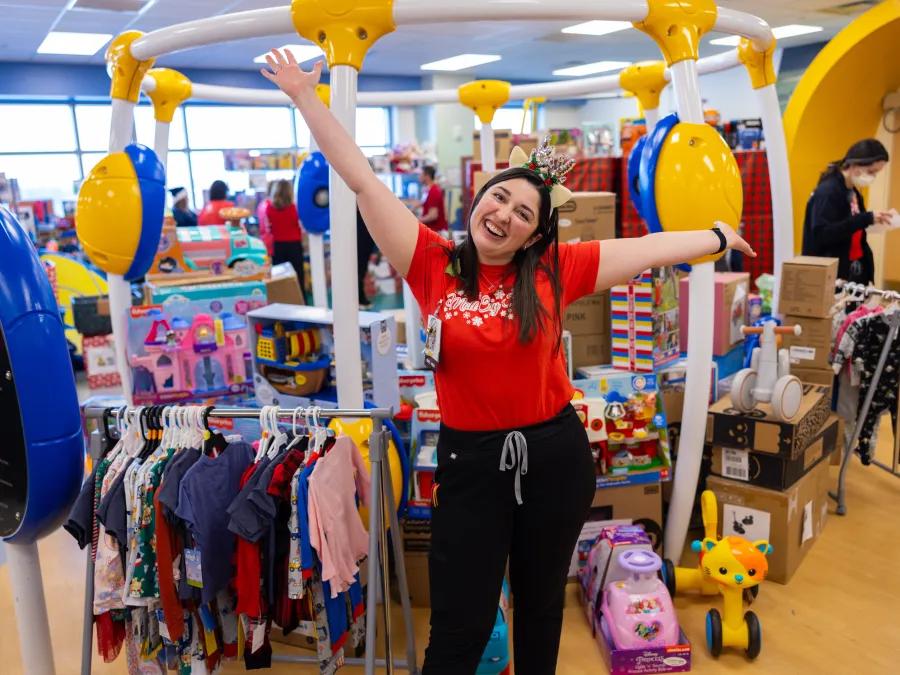 A staff or volunteer in a red t-shirt stands with her arms extended excitedly in front of a room full of kids' clothing and toys.