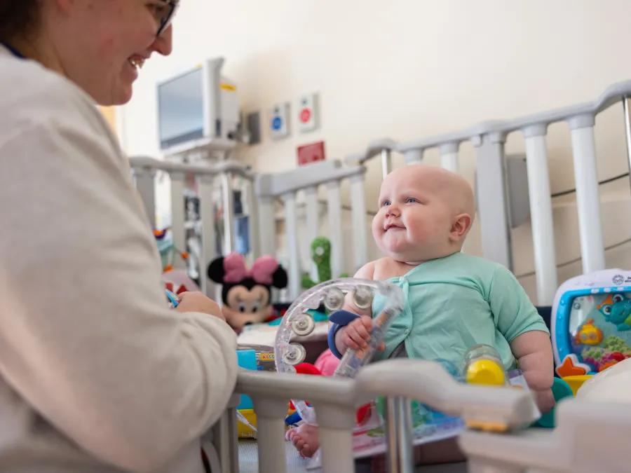 Smiling baby in white crib filled with toys with hospital monitor in background and female medical professional looking on