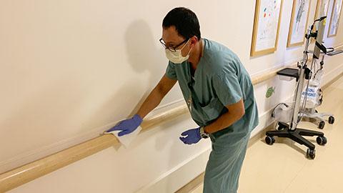 Man in scrubs, surgical mask, and gloves wiping down the rainling the the hospital hallway