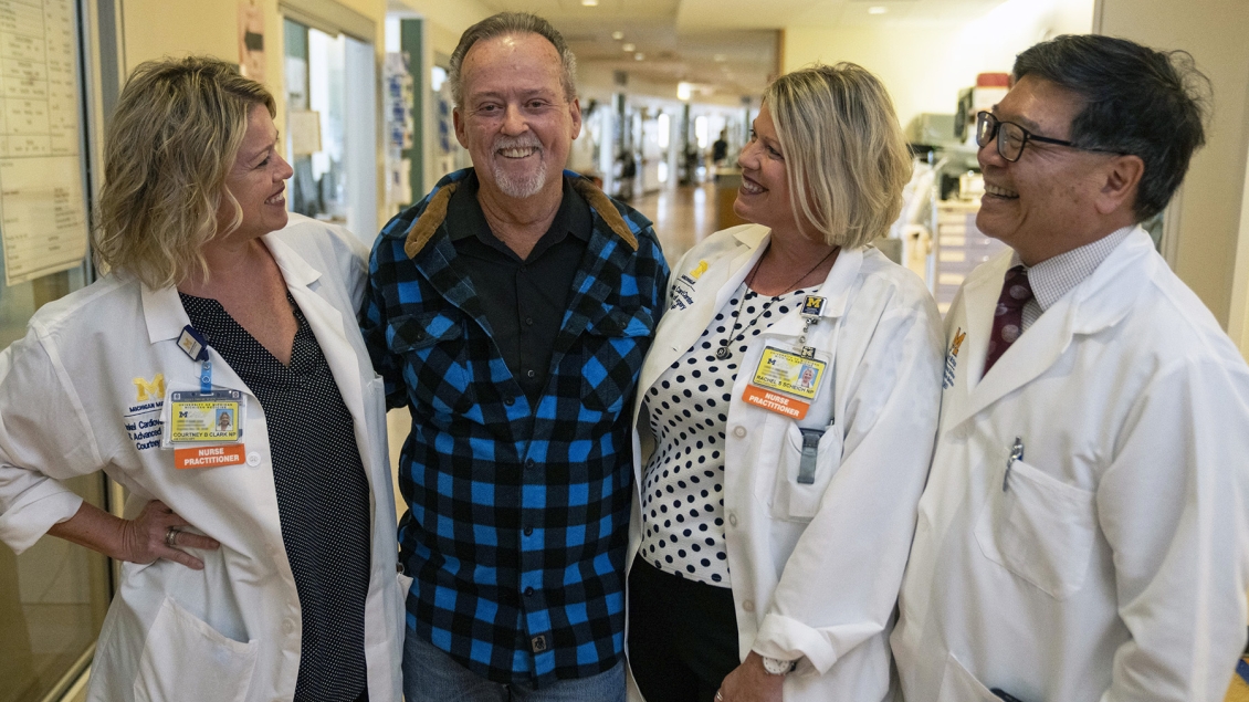 A smiling patient stands in a hospital hallway with three members of his care team in white coats. The group is standing close together, smiling warmly, showing a supportive relationship.