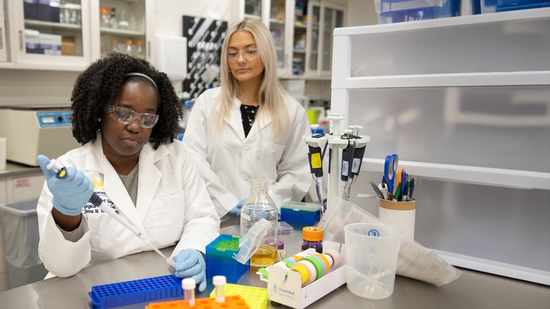 Researchers in the Newman Lab. One with a pipette and the other watching.