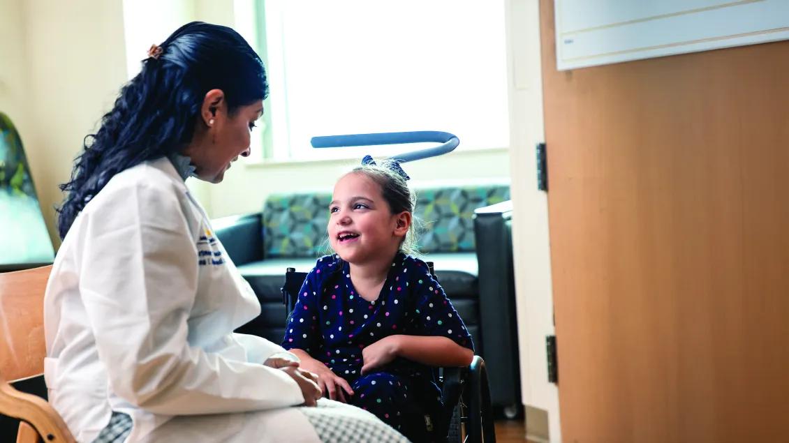 Little girl in wheelchair wearing polka-dot dress smiling and talking with seated dark-haired female doctor in white coat