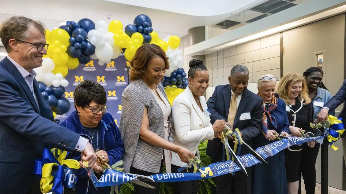 Group of people cutting ribbon for U-M Health Ypsilanti Health Center opening with maize and blue balloons in background