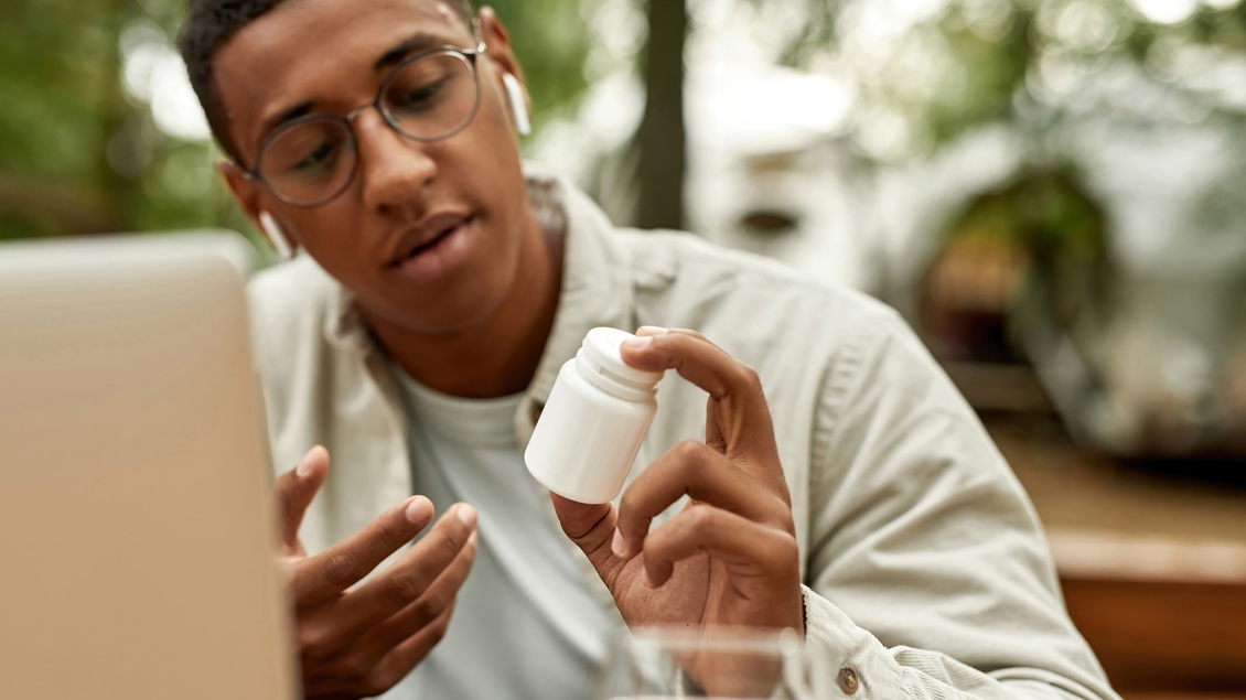 Young man in front of a laptop and with earbuds in his ears looking a white prescription bottle