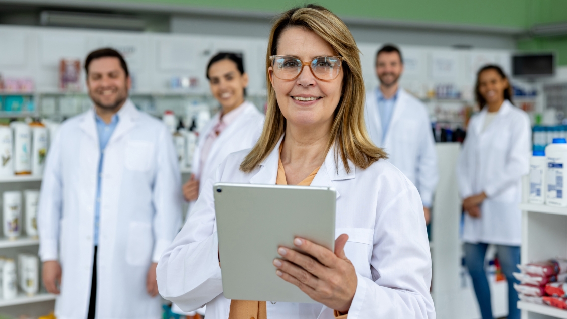 Pharmacist holding a tablet in a pharmacy with team members standing behind her.