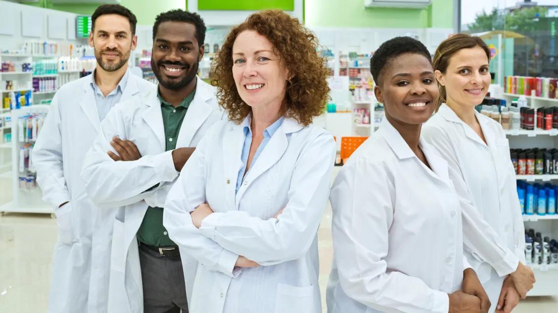 Group of healthcare professionals in lab coats smiling and posing for a photo inside a pharmacy.