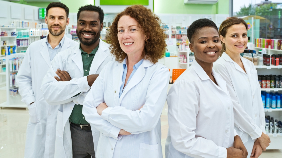 Group of healthcare professionals in lab coats smiling and posing for a photo inside a pharmacy.