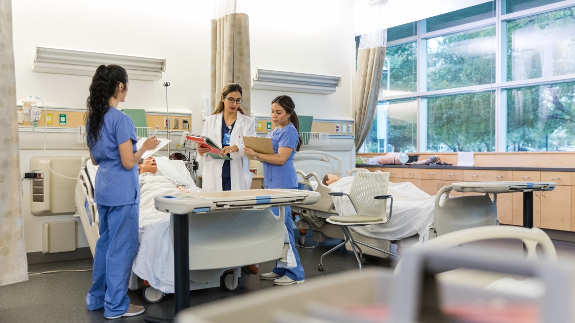 The female hospital trainer stands with the two interns in the medical classroom and repeats instructions so they can confirm their notes are correct.