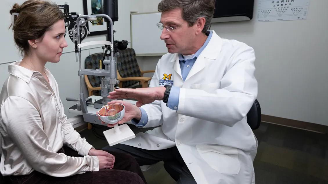 Grant Comer, MD in an exam room holding a replica of an eye as he is explaining an eye disease to a patient sitting in front of him. The patient is wearing a white blouse and dark slacks.