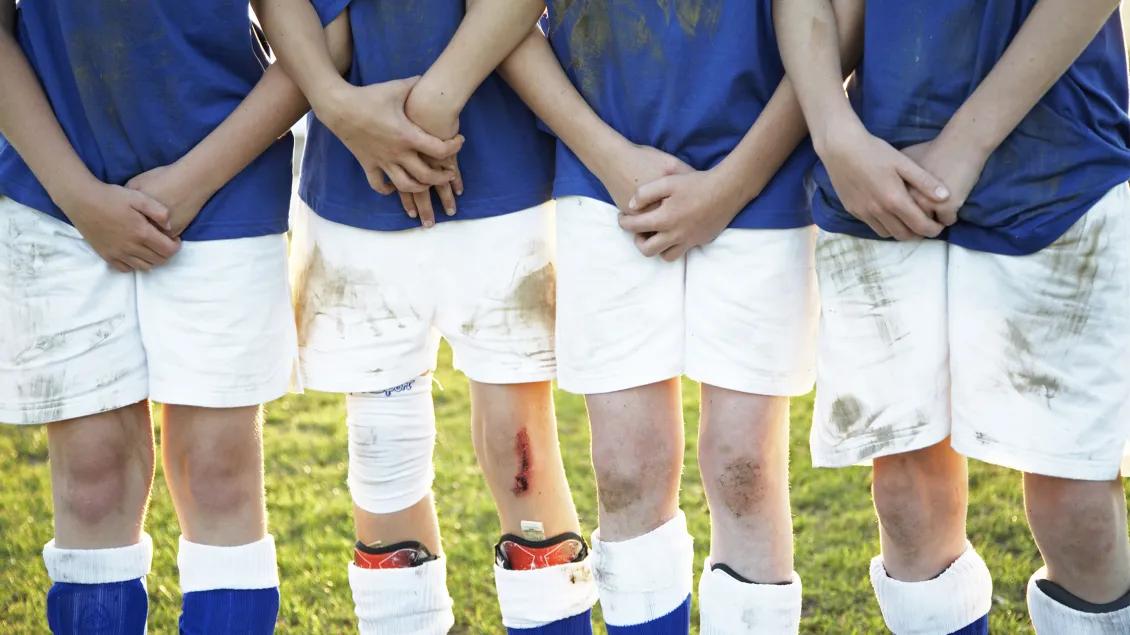 Row of kids with skinned knees shown from elbows down wearing blue shirts and dirty white shorts