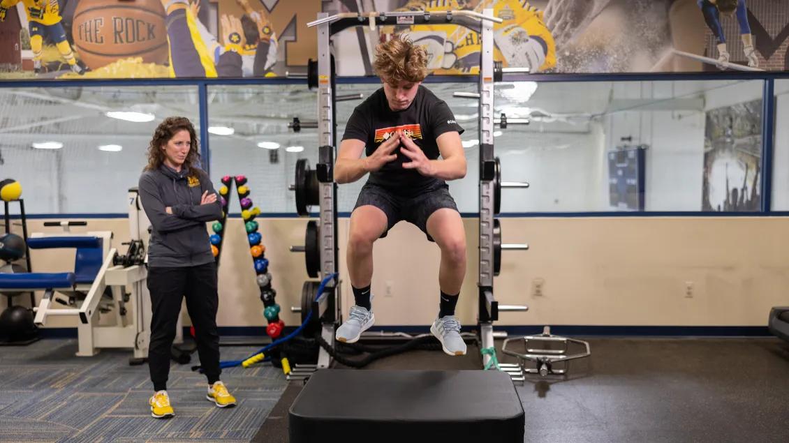 Young male athlete in mid-jump on training equipment with trainer standing by