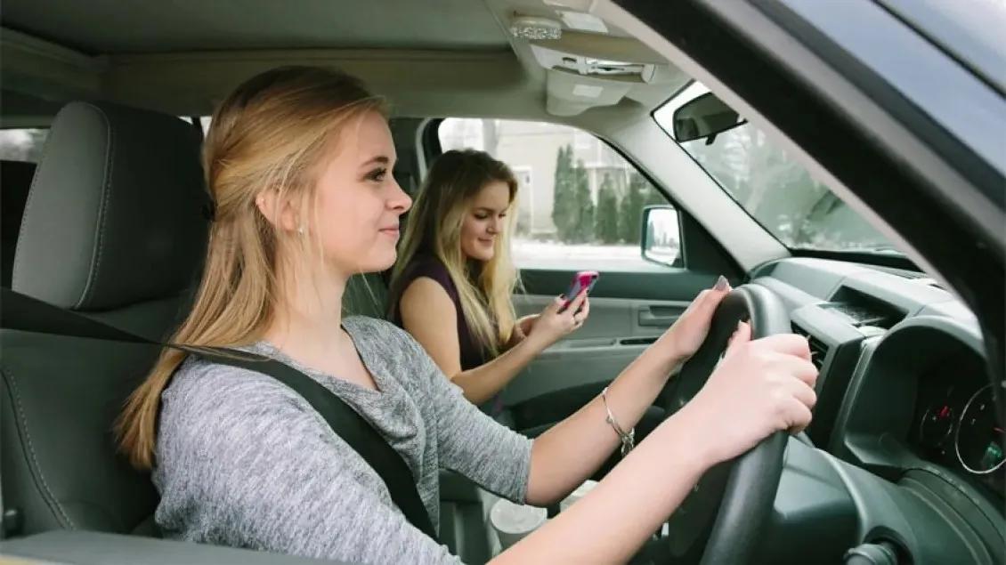 Teen driver sitting in the driver's seat with hands on the steering wheel while a friend in the front passenger seat looking at her phone.