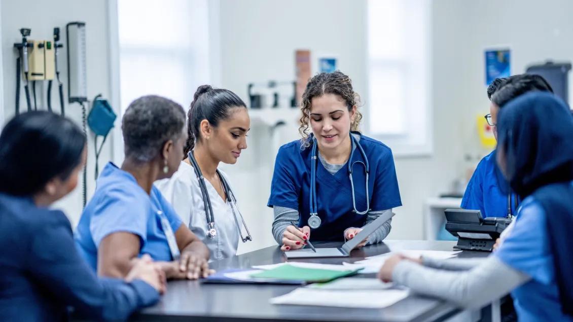 A small group of medical professionals, led by a doctor, are participating in team training as they sit around a table together. They are each dressed professionally and are focused on the meeting.
