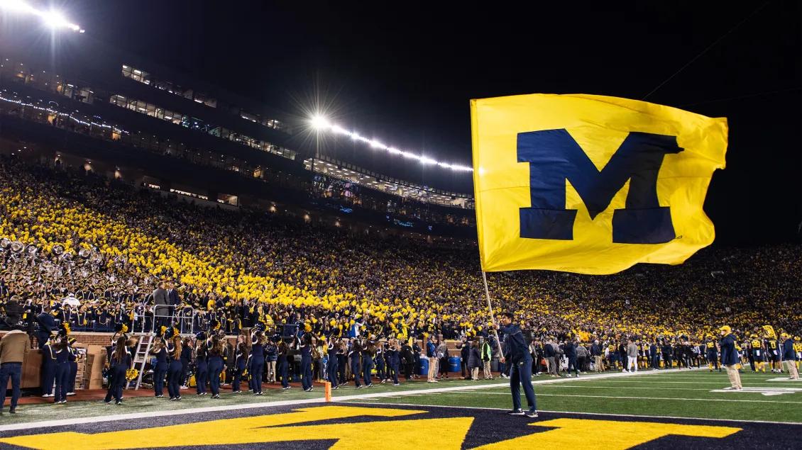 Man holding huge maize flag with blue block M at U-M football stadium with crowd in background