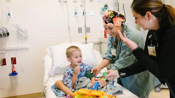 Little boy wearing children's hospital gown in hospital bed with toys and two female medical professionals engaging with him