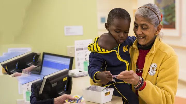 Little boy Jonathon from Uganda wearing blue M jacket, being given sticker by brown-skinned woman in maize jacket