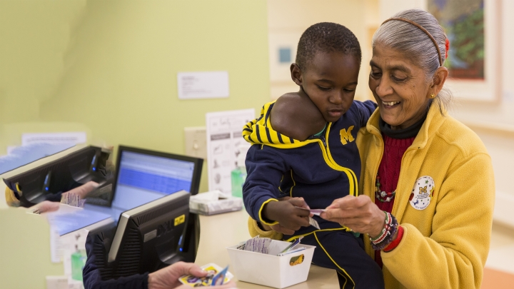 Little boy Jonathon from Uganda wearing blue M jacket, being given sticker by brown-skinned woman in maize jacket