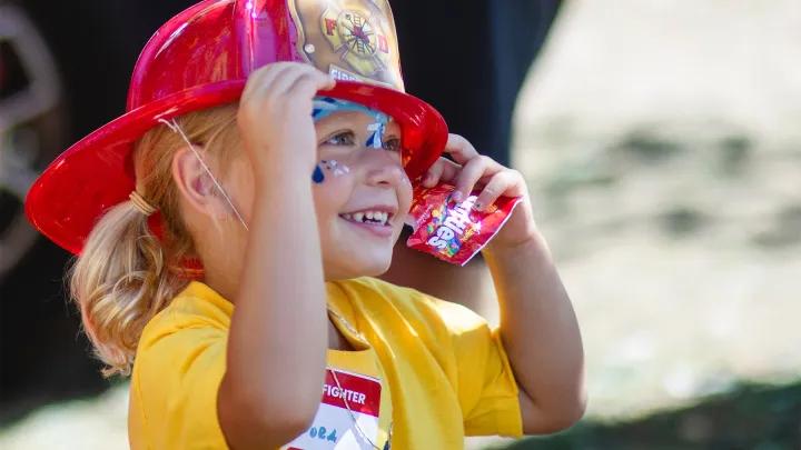 Little girl in yellow shirt with block M wearing red plastic fire hat with pack of Skittles in her hand