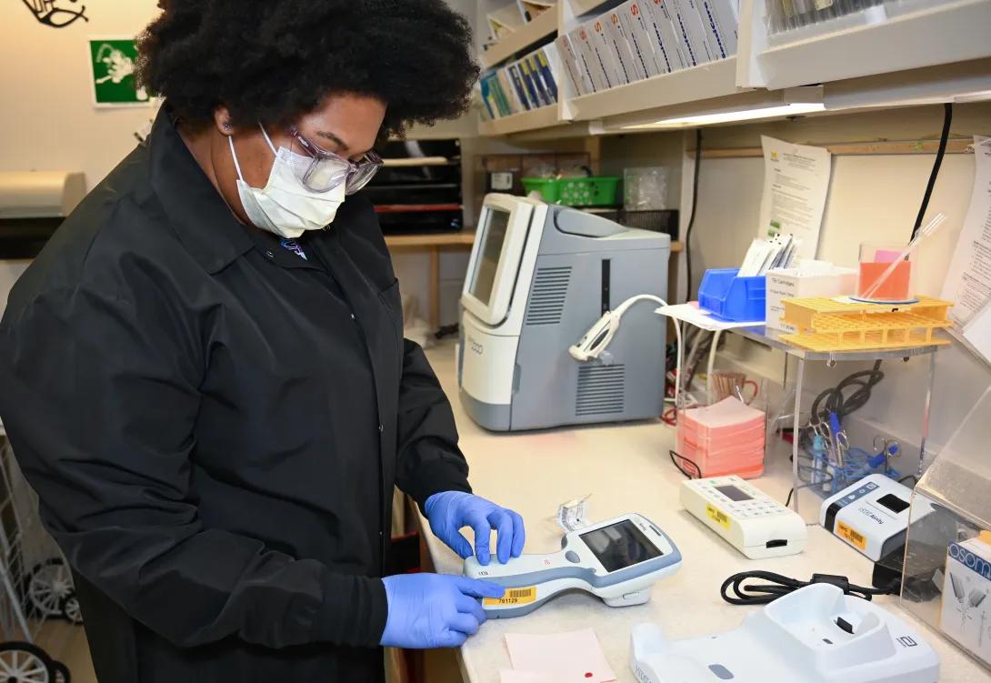 A lab technician wearing a mask, gloves, and protective coat operates a handheld medical testing device at a laboratory workstation.