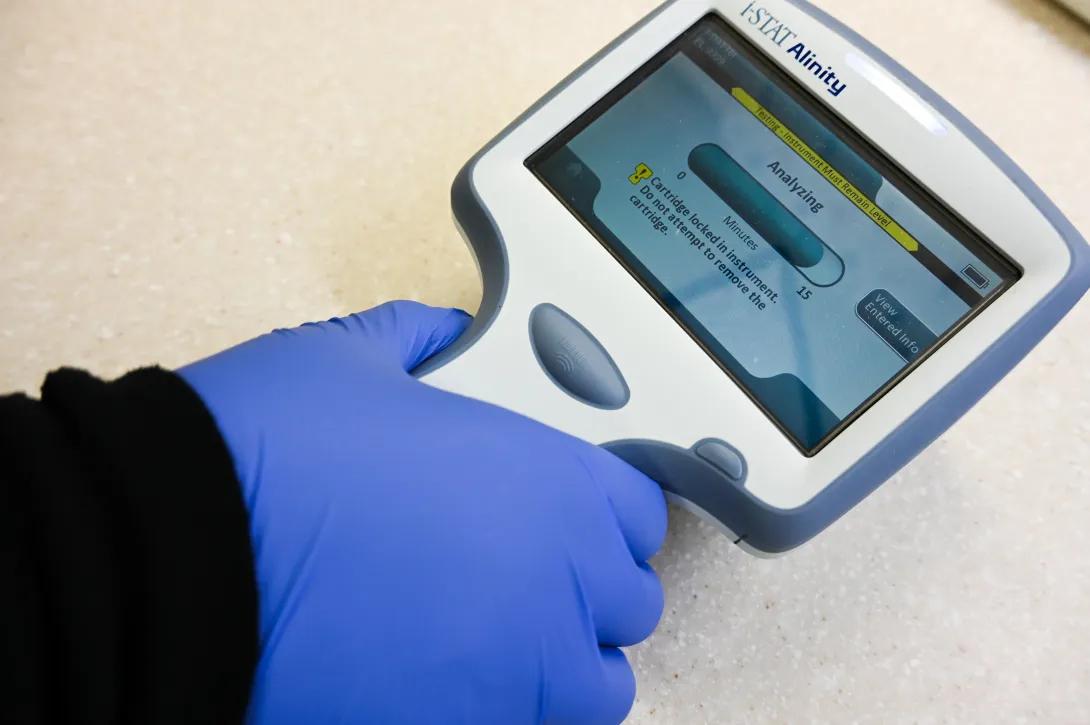 Close-up of a gloved hand holding a handheld diagnostic device labeled “i-STAT Alinity,” with the screen displaying an “Analyzing” status and test progress information on a lab countertop.