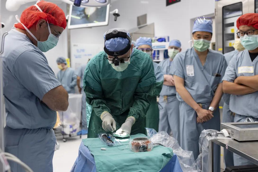 A group of medical professionals in surgical attire are gathered in an operating room. One individual is actively engaged over a table covered with sterile blue drapes. Others watch attentively. The environment is equipped with medical instruments and equipment, creating a typical surgical setting.