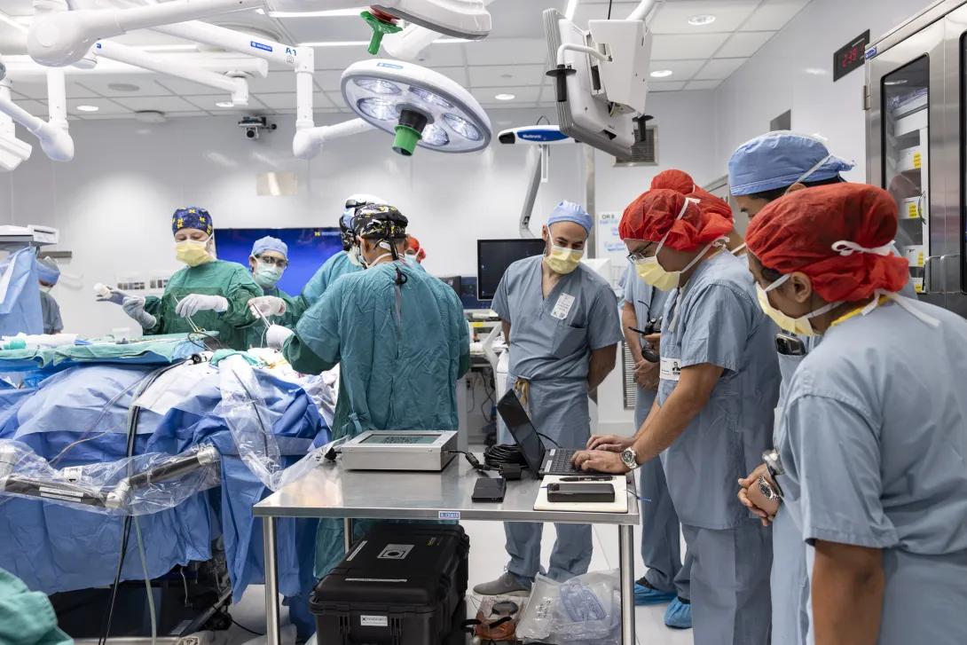 A team of medical professionals in an operating room performs a surgical procedure. The room is equipped with modern medical equipment, including overhead lights and multiple monitors. Some team members are dressed in teal scrubs and others in blue. Another group observes closely.