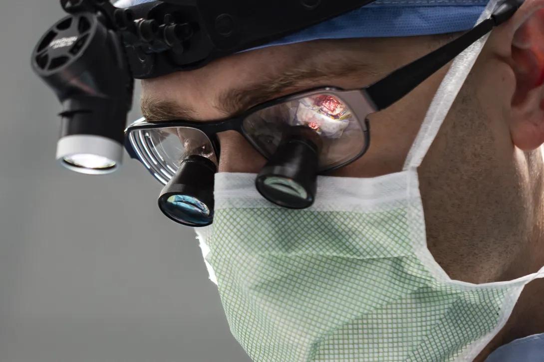 A person wearing magnifying surgical glasses, a headlamp, and a surgical mask is focused on performing a procedure. The background is blurred, emphasizing the concentration on the task.