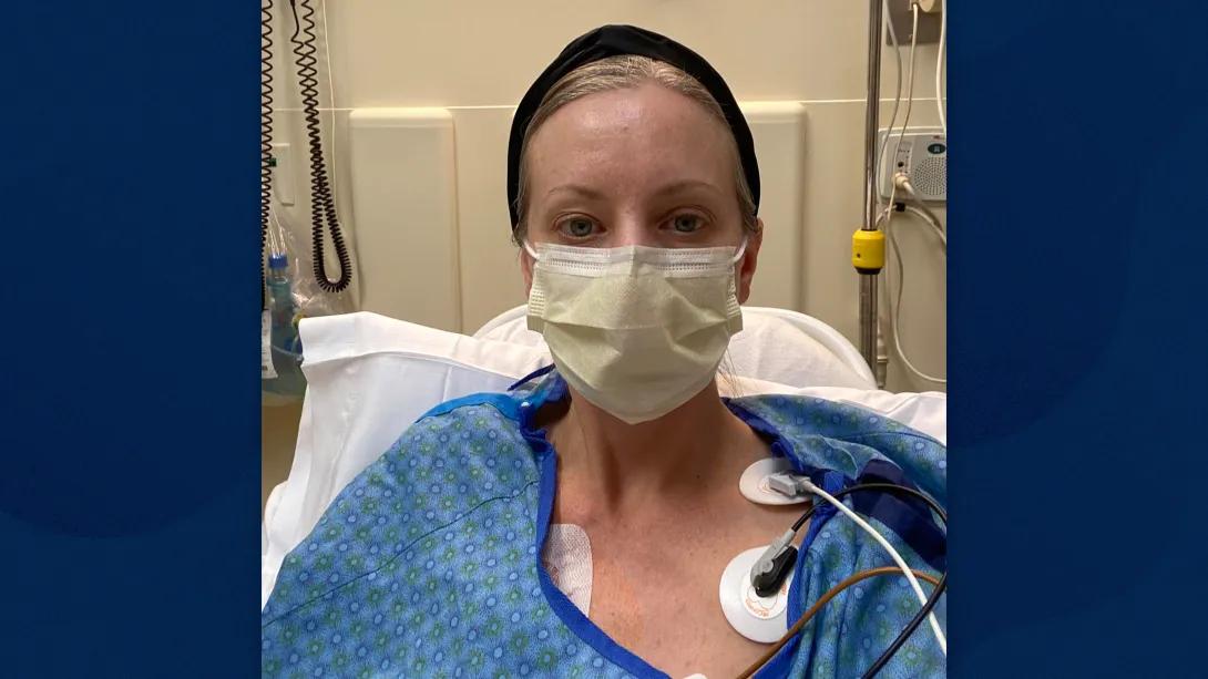 Woman in blue hospital gown, sitting in hospital bed with face mask and cap. Visible chest monitors are stuck with adhesive on her body.